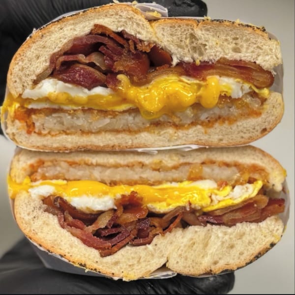 A sliced bagel sandwich from Emma's Deli, filled with bacon, fried eggs, and hash browns.