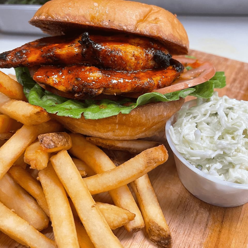 Emma's Deli's grilled chicken sandwich with lettuce, tomato, fries, and coleslaw on a wooden board.