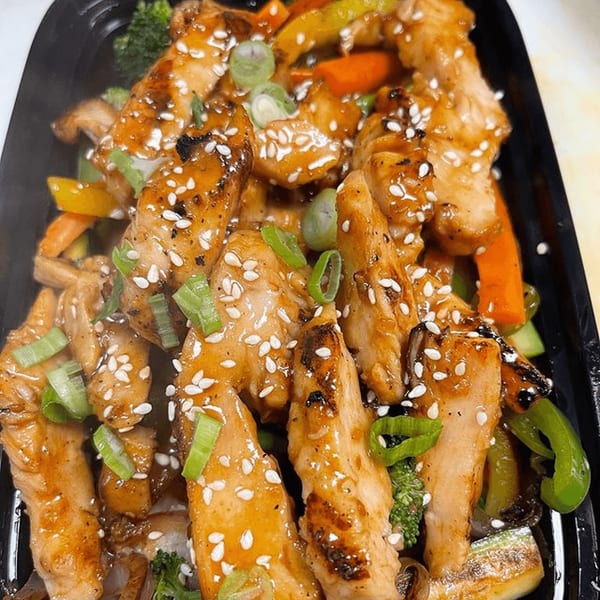 Teriyaki chicken strips with sesame seeds, green onions, and mixed vegetables in a black container.