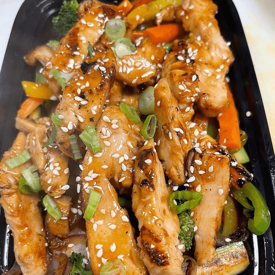 Teriyaki chicken strips with sesame seeds, green onions, and mixed vegetables in a black container.