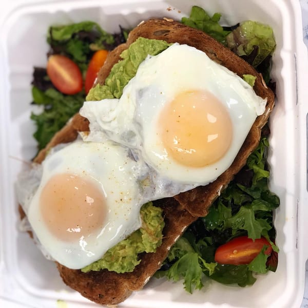 Avocado toast with two fried eggs and a side salad with cherry tomatoes in a takeout container.