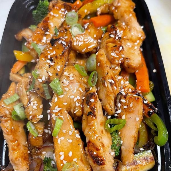 Teriyaki chicken strips with sesame seeds, green onions, and mixed vegetables in a black container.