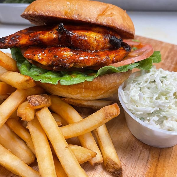 Emma's Deli's grilled chicken sandwich with lettuce, tomato, fries, and coleslaw on a wooden board.